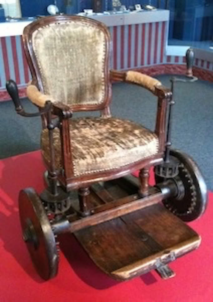 Fauteuil roulant ancien en bois avec accoudoirs rembourrés et dossier en tissu, posé sur un tapis rouge.