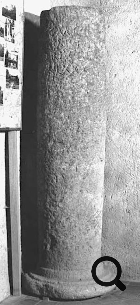 Colonne en pierre avec des motifs sculptés, posée sur une base circulaire, à côté d'un mur.