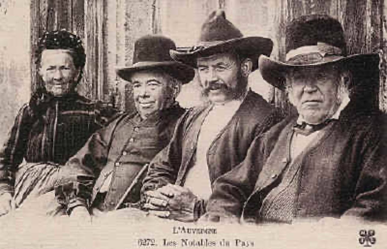 Quatre personnes assises portant des chapeaux et des vêtements anciens, posant devant un fond en bois.
