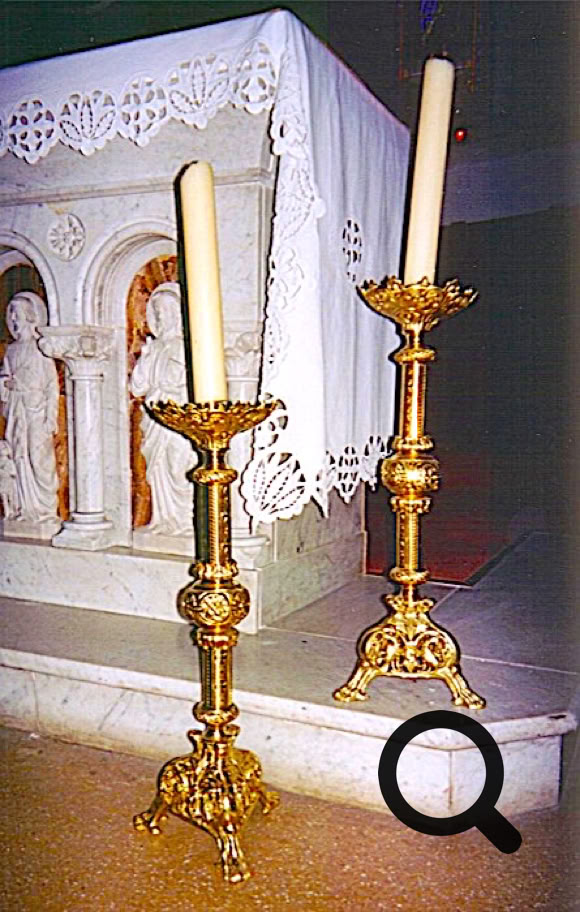 Deux chandeliers dorés avec des bougies allumées devant un autel en pierre décoré de motifs sculptés et recouvert d'une nappe blanche.
