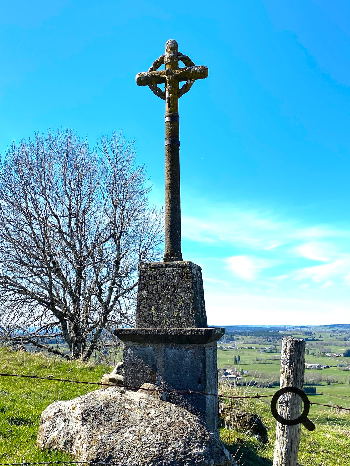 Croix en pierre sur un socle, avec un arbre sans feuilles en arrière-plan et un paysage de campagne sous un ciel bleu.