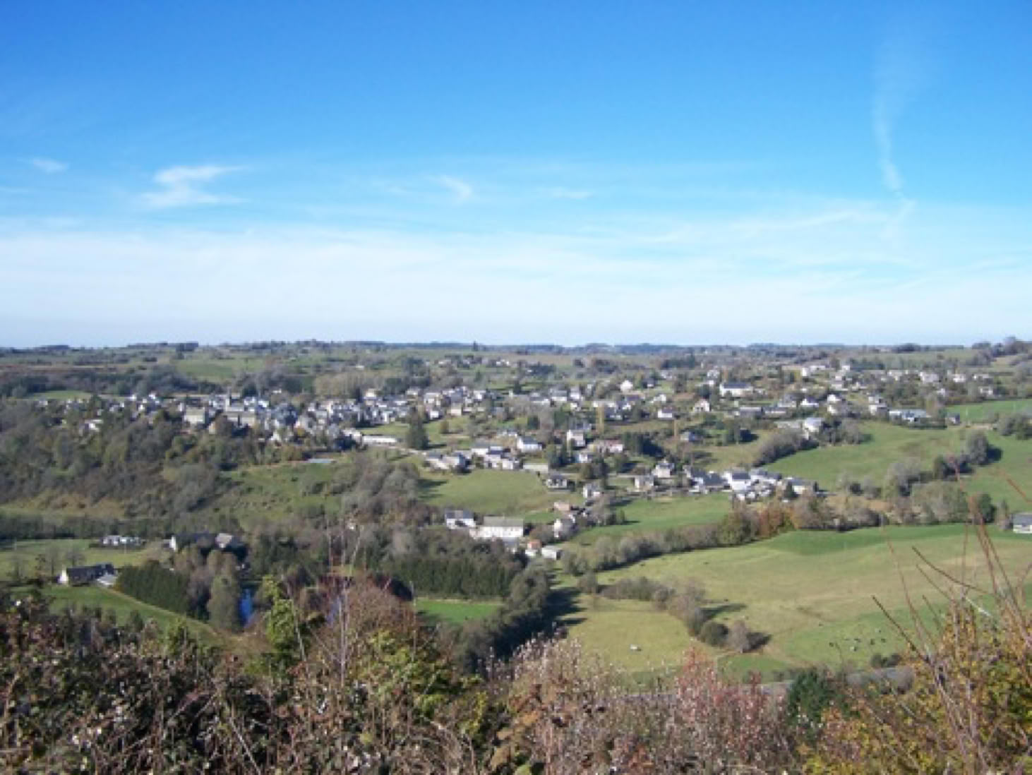 Vue panoramique d'un paysage rural avec des collines verdoyantes, des champs, et un village avec des maisons dispersées sous un ciel bleu.