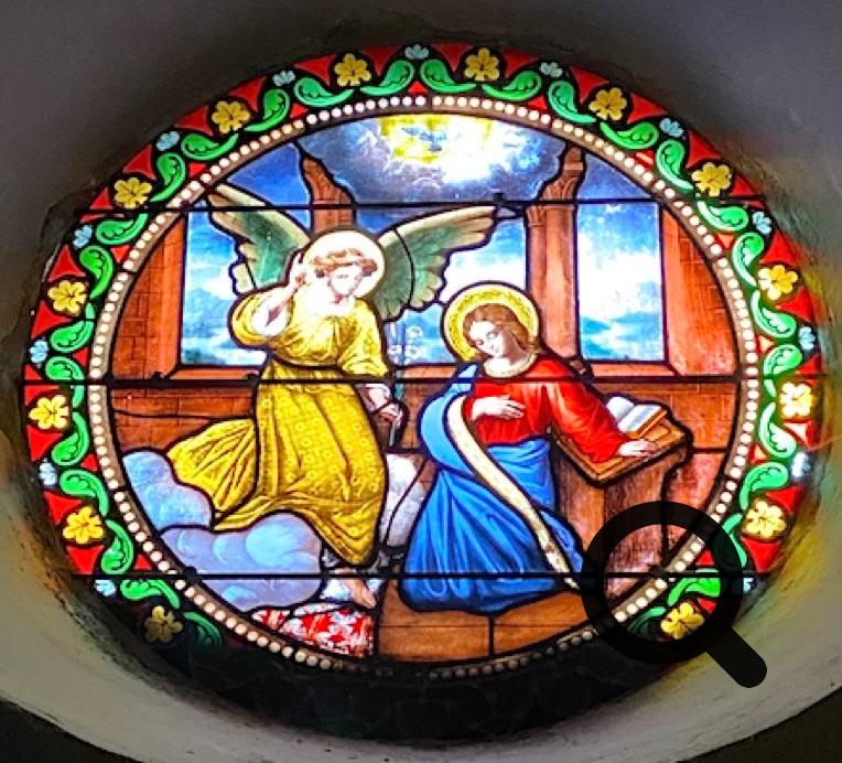 Vitrail circulaire représentant un ange avec des ailes vertes parlant à une femme agenouillée, entouré d'un motif floral coloré.