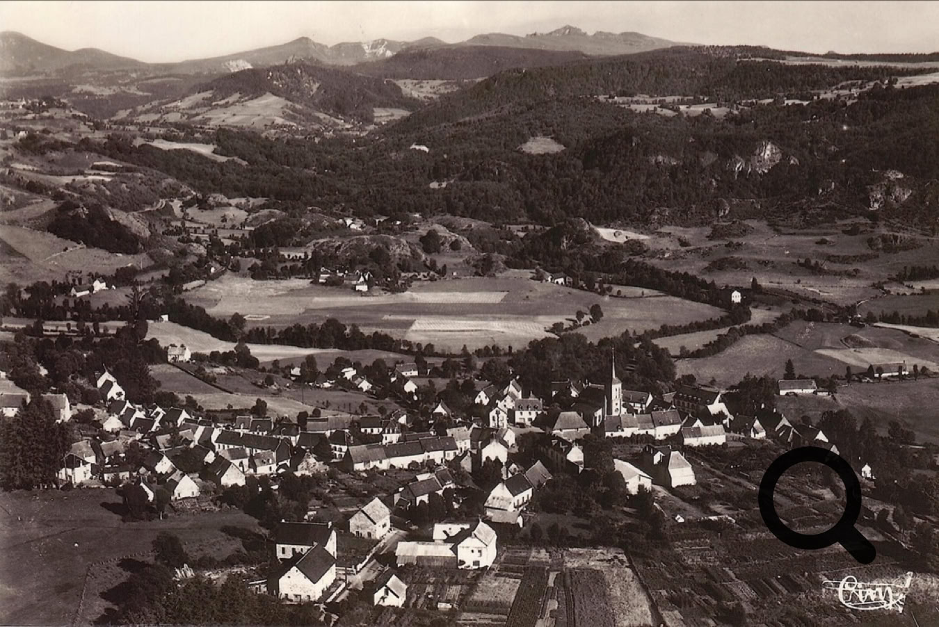 Vue aérienne d'un village avec des maisons dispersées, entouré de collines et de montagnes à l'arrière-plan.