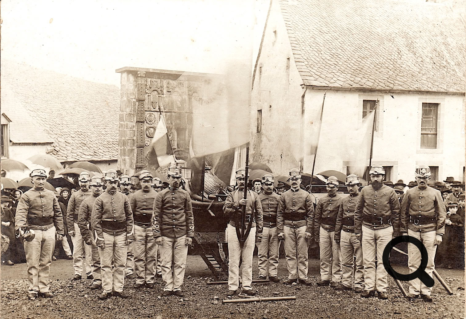 Groupe de sapeurs-pompiers en uniforme posant devant des bâtiments anciens avec un drapeau visible à l'arrière-plan.