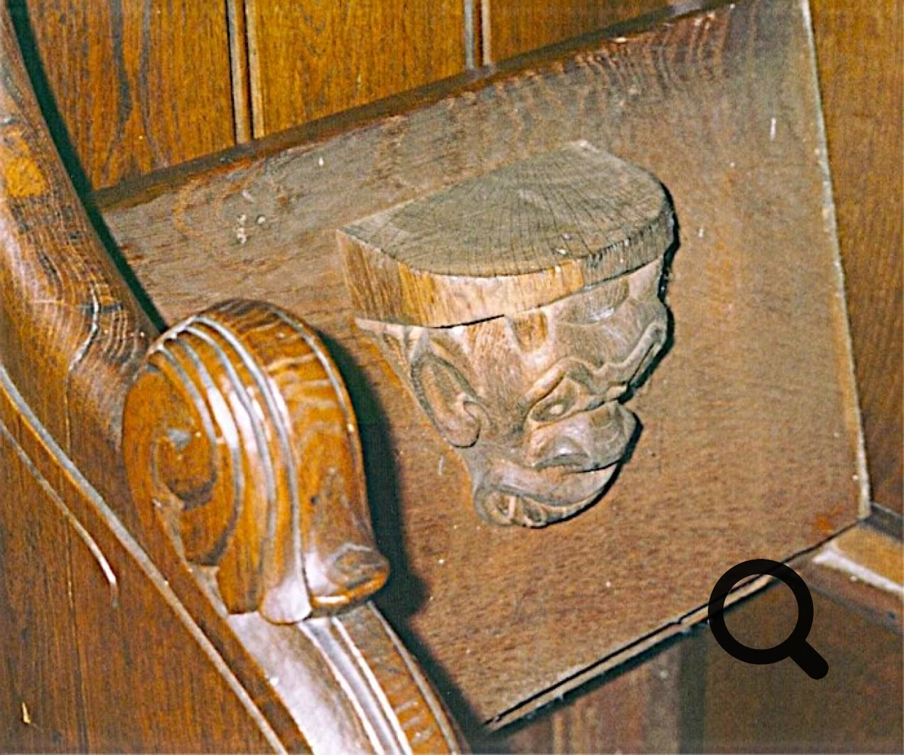 Détail d'un banc en bois avec une sculpture en forme de visage grotesque.