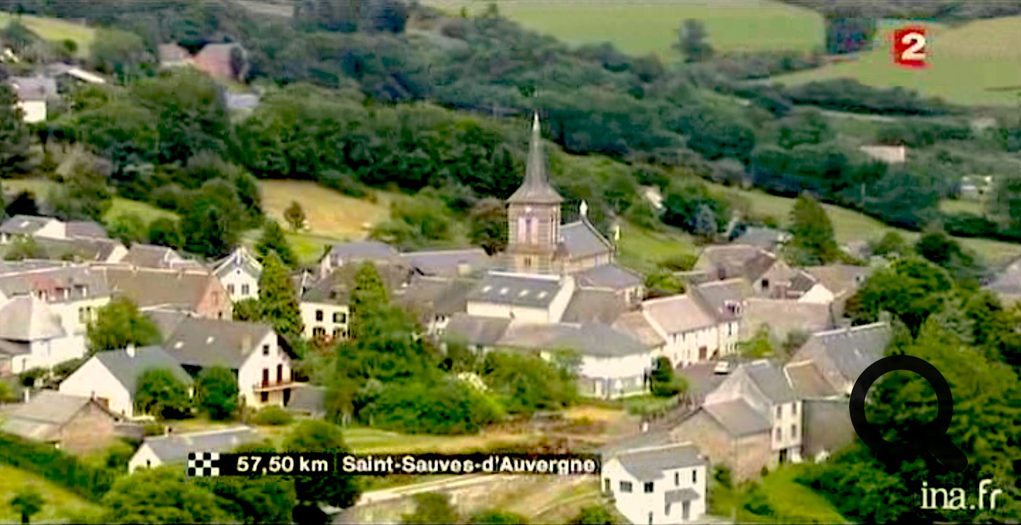 Vue aérienne d'un village avec des maisons, une église au centre, entouré de verdure et de collines.