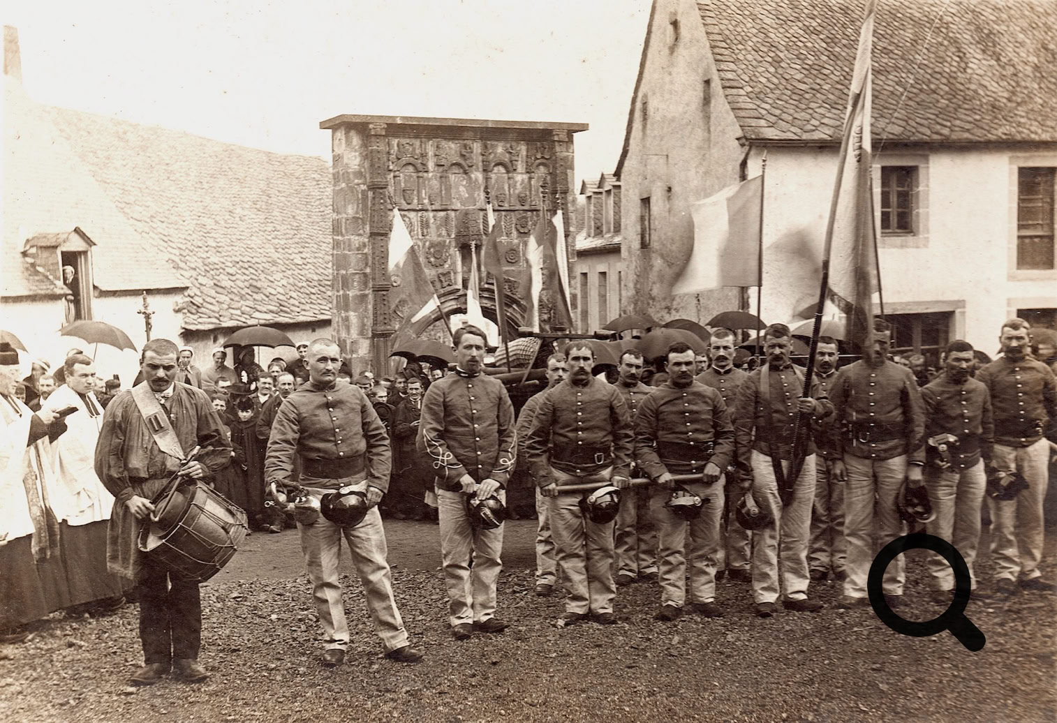 Groupe de sapeurs-pompiers en uniforme alignés devant des bâtiments, avec des drapeaux et des personnes en arrière-plan.