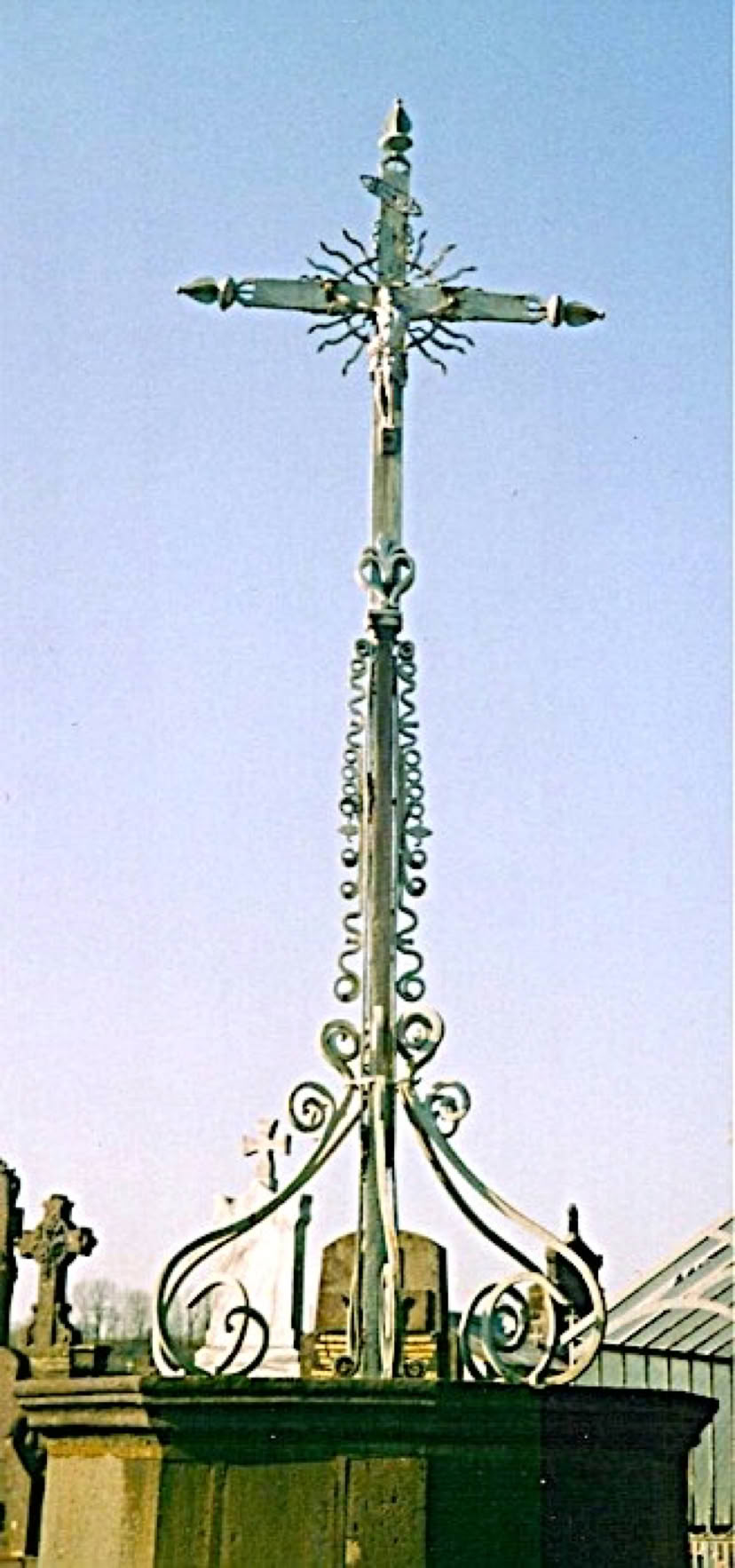 Croix en métal ornée de motifs décoratifs, sur un toit avec ciel bleu en arrière-plan.