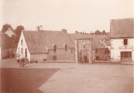 Une place de village avec des bâtiments anciens, dont une maison à toit de chaume et une autre avec une façade en pierre. Deux personnes marchent à gauche.
