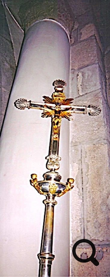 Croix dorée ornée devant une colonne blanche et un mur en pierre.
