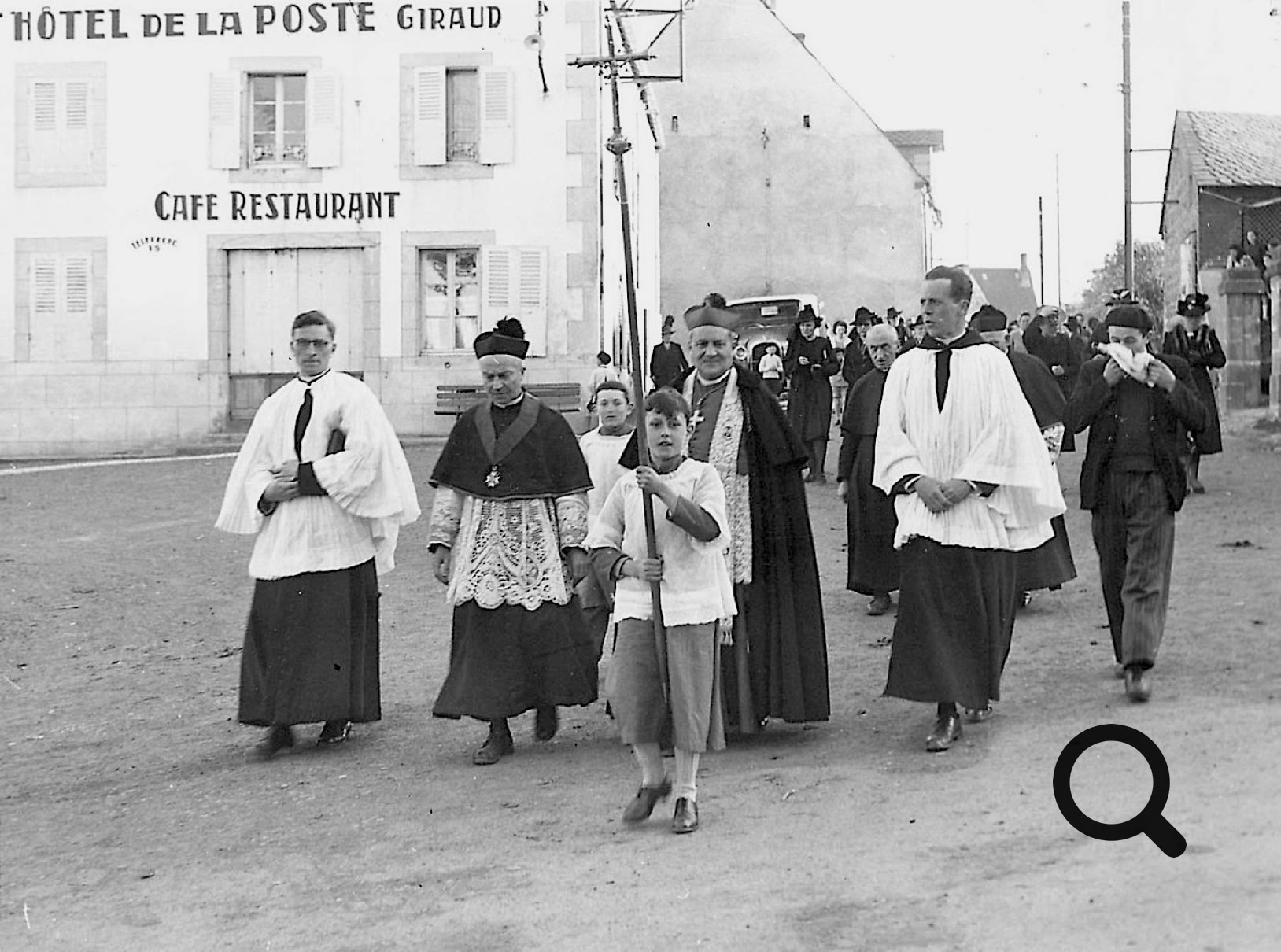 Un groupe de personnes en vêtements religieux marche dans une rue, avec un bâtiment marqué "Hôtel de la Poste" et "Café Restaurant" en arrière-plan.