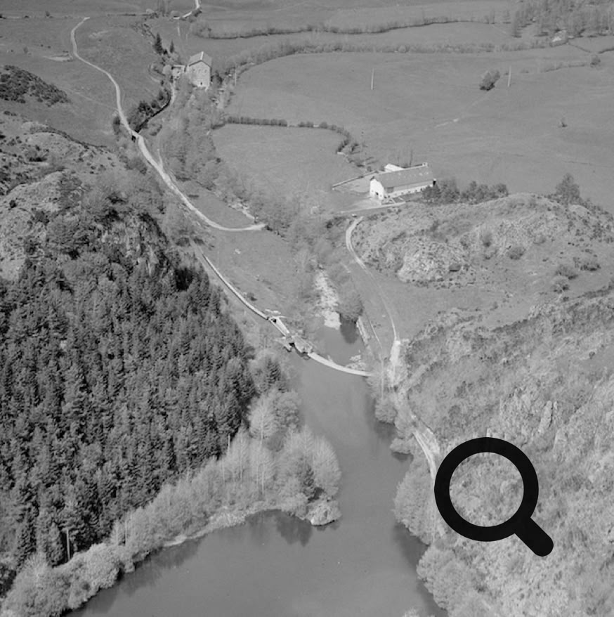 Vue aérienne en noir et blanc d'un paysage rural avec une rivière sinueuse, des collines boisées, des champs et quelques bâtiments dispersés.